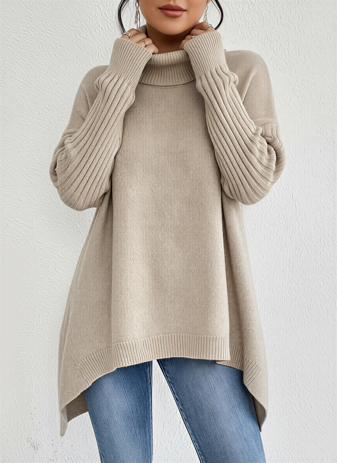 Femme portant un pull à col roulé beige à la coupe ample et fluide. Le vêtement présente des manches longues côtelées et un ourlet asymétrique, créant une allure moderne et élégante. Associé à un jean ajusté, l’ensemble offre un style confortable et chic, idéal pour la saison automne-hiver. La texture douce du tricot met en valeur la qualité et le raffinement du tissu.