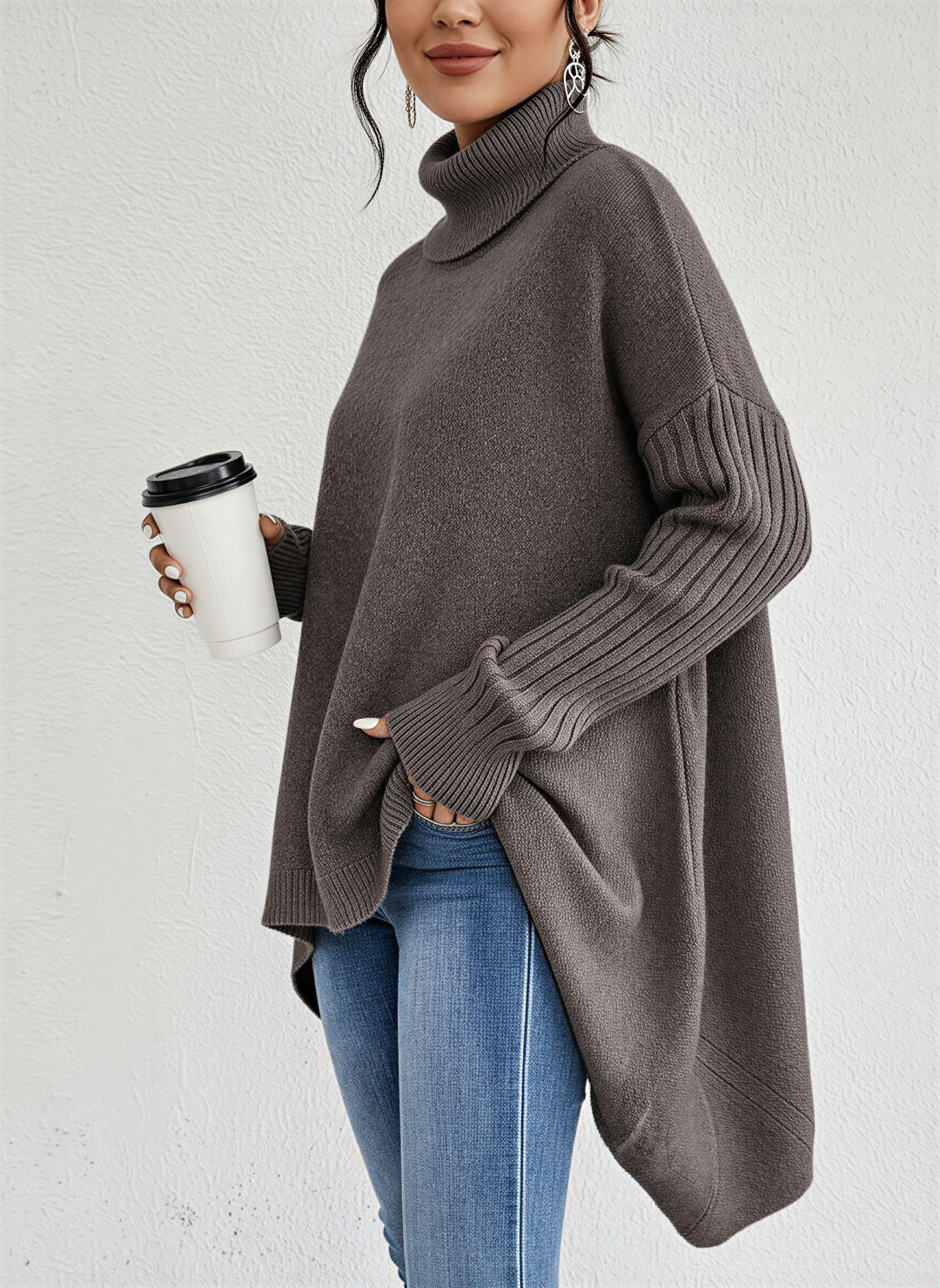 Femme portant un pull à col roulé ample de couleur taupe, à manches longues côtelées. Le vêtement présente une coupe fluide avec un ourlet asymétrique, apportant une touche moderne et élégante. Porté avec un jean ajusté, l’ensemble crée un style à la fois chic et décontracté, idéal pour les journées d’automne. La femme tient un gobelet de café à la main, ajoutant une note cosy et urbaine à la tenue.