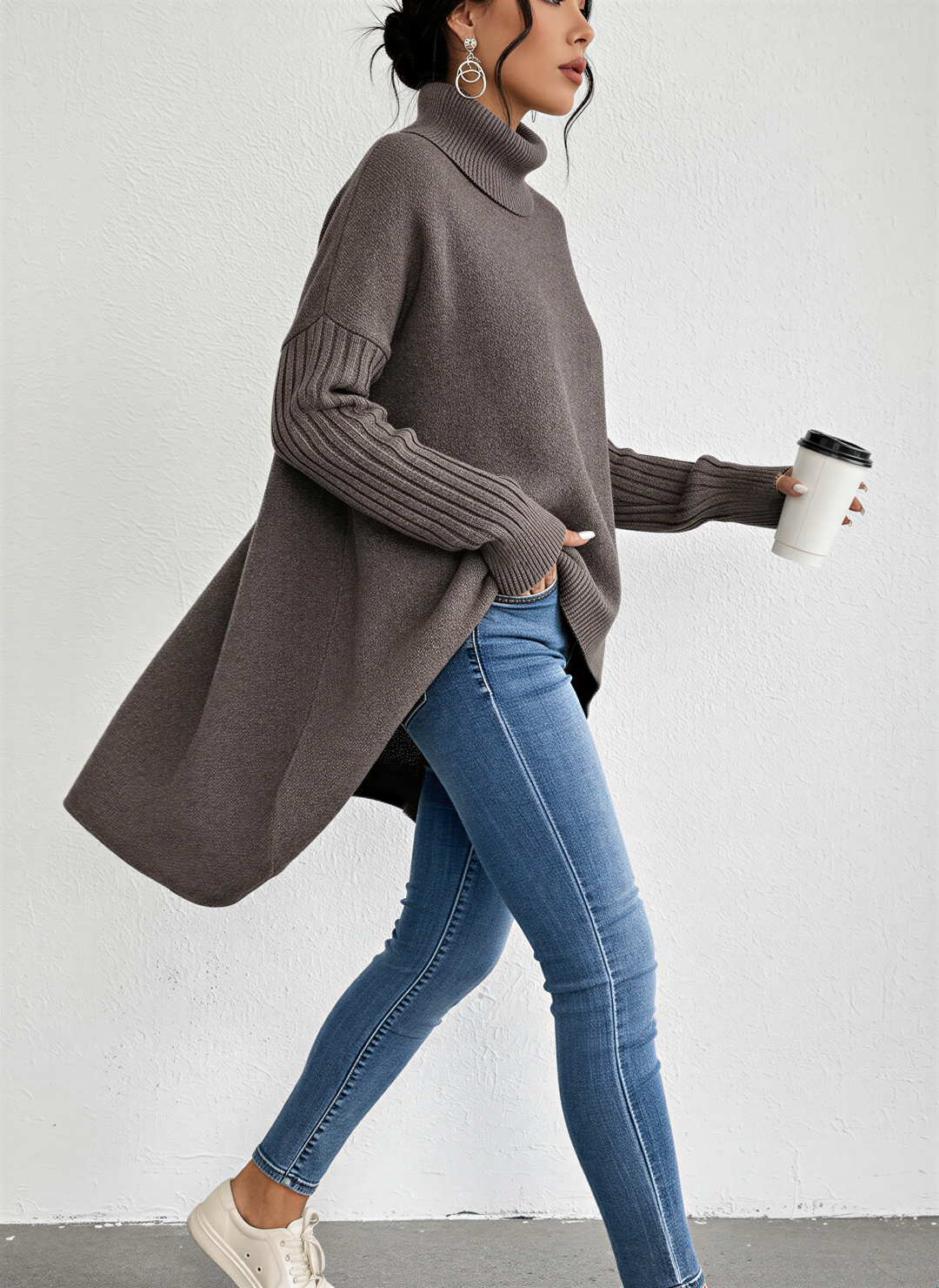 Femme marchant avec élégance, vêtue d’un pull à col roulé marron à la coupe ample et moderne. Le vêtement présente des manches longues côtelées et un ourlet asymétrique plus long à l’arrière, offrant un tombé fluide et raffiné. Porté avec un jean slim bleu et des baskets blanches, l’ensemble crée un look chic et décontracté, parfait pour les journées d’automne ou d’hiver. La tasse de café à la main ajoute une touche urbaine et contemporaine à la scène.
