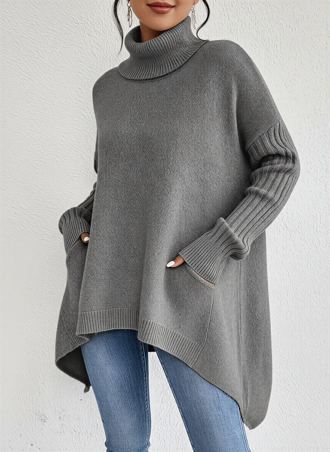 Femme portant un pull à col roulé gris à la coupe ample et élégante. Le vêtement se distingue par ses manches longues côtelées et son ourlet asymétrique, offrant une allure moderne et raffinée. Les poches discrètes sur les côtés ajoutent une touche pratique et décontractée. Associé à un jean ajusté, cet ensemble crée un style chic et confortable, parfait pour la saison froide.