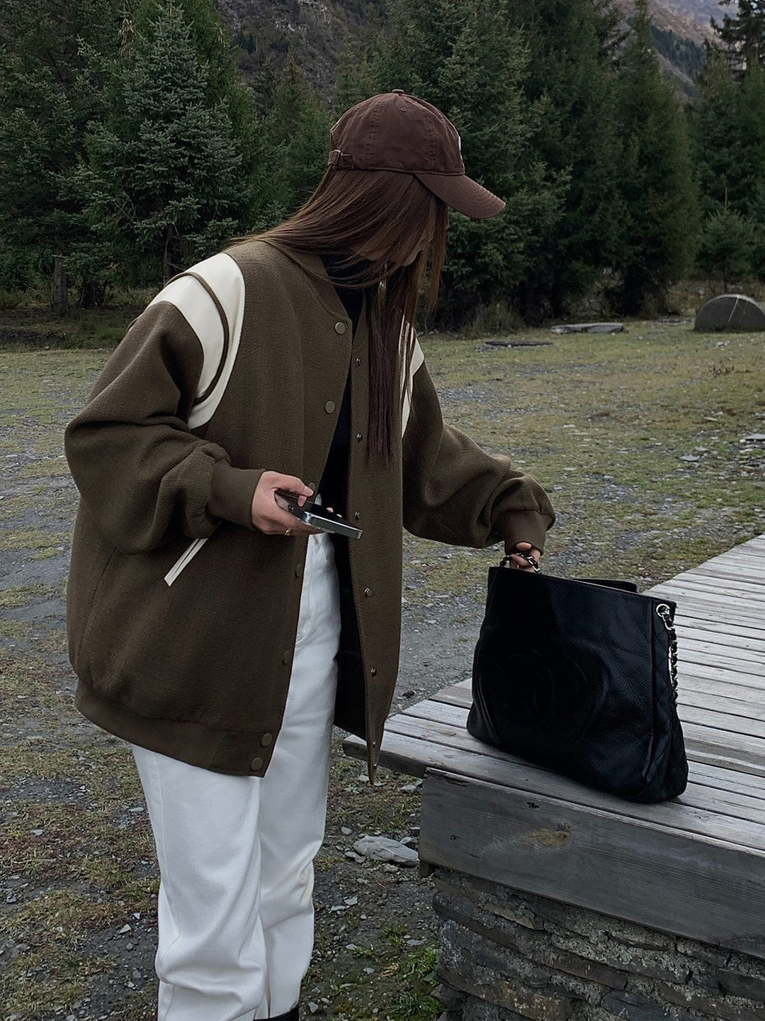 veste coupe droite couleur brun avec empiècements crème sur les épaules, fermeture à boutons, associée à un pantalon blanc et un sac noir, look tendance en extérieur dans un cadre nature.