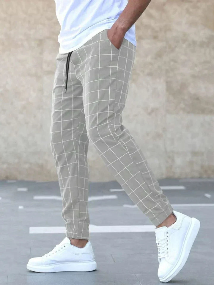 Homme portant le pantalon classique Juste pour hommes, beige à carreaux, coupe ajustée et chevilles resserrées, avec poches latérales et cordon de serrage, associé à un t-shirt blanc et des baskets blanches  style tendance et élégant.