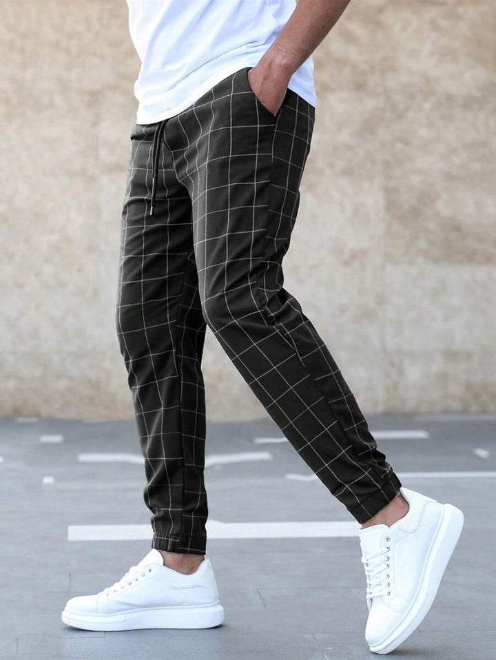 Homme portant le pantalon classique Juste pour hommes, noir à carreaux blancs, coupe slim avec taille élastique et cordon de serrage, chevilles resserrées et poches latérales, porté avec un t-shirt blanc et des baskets blanches look tendance et élégant.