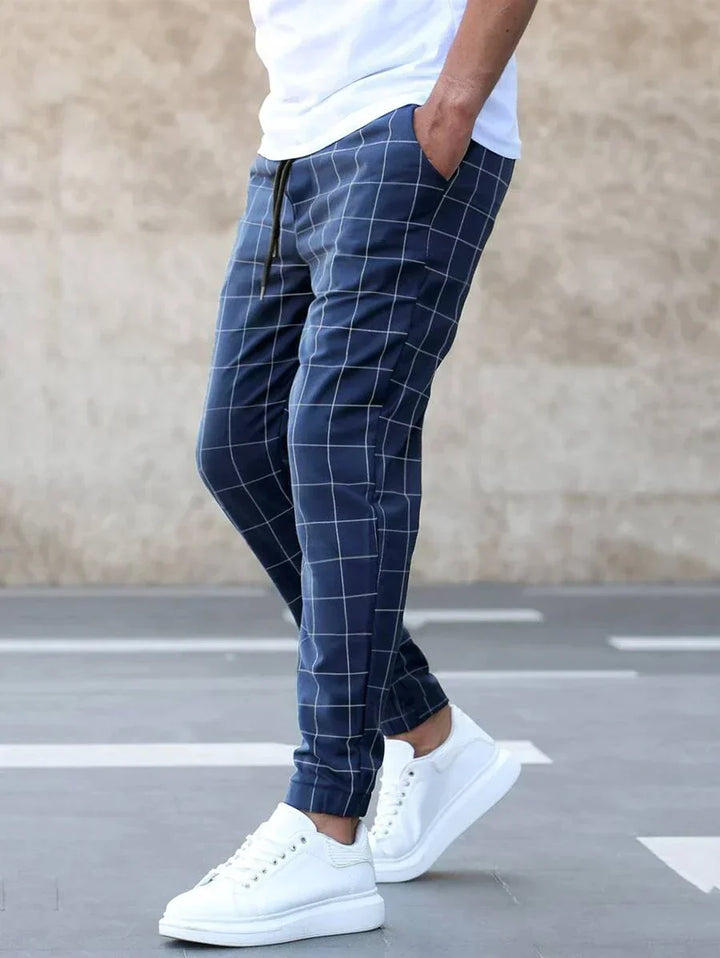 Homme portant le pantalon classique Juste pour hommes, bleu marine à carreaux, coupe slim avec taille élastique et cordon de serrage, chevilles resserrées et poches latérales, associé à un t-shirt blanc et des baskets blanches style tendance et chic.