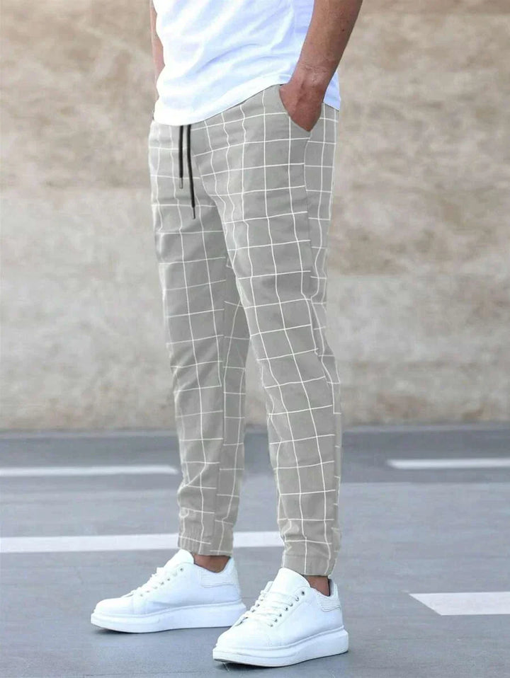 Homme portant le pantalon classique Juste pour hommes, beige à carreaux blancs, coupe ajustée avec taille élastique et cordon de serrage, chevilles resserrées et poches latérales, porté avec un t-shirt blanc et des baskets blanches  look tendance et confortable.