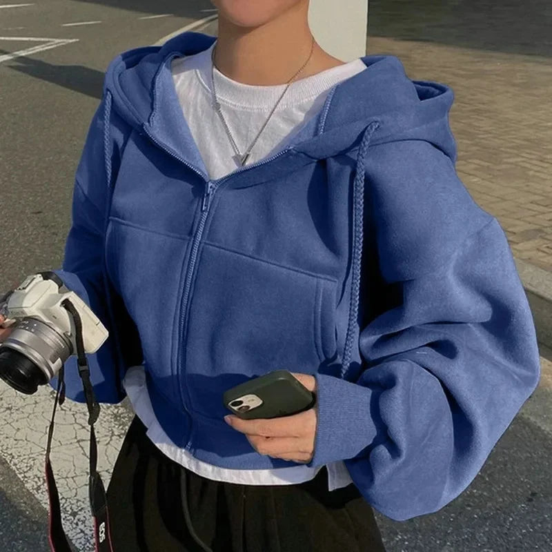 Une femme porte un sweat à capuche bleu clair, ajusté avec une fermeture éclair. Elle tient un appareil photo dans une main et un téléphone portable dans l'autre. Elle se tient à l'extérieur, avec un fond simple et urbain, sous un ciel ensoleillé.