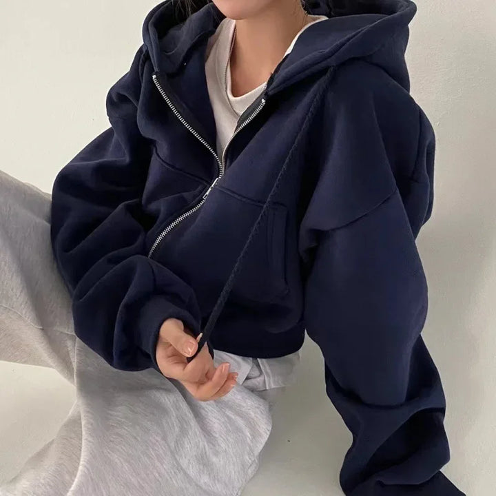 Une femme porte un sweat à capuche bleu marine avec une coupe ample et décontractée. Elle est assise, portant un pantalon gris. Elle tire sur le cordon de serrage du sweat et son visage n'est pas visible. L'arrière-plan est simple, avec des tons neutres.
