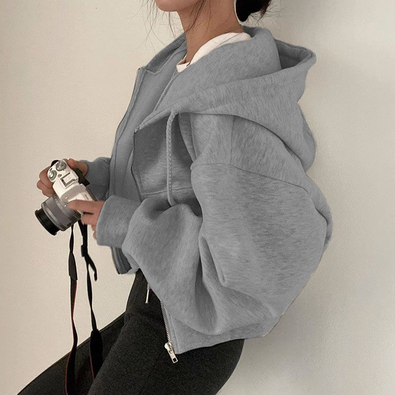 Une femme porte un sweat à capuche gris, avec des manches amples et un style décontracté. Elle tient un appareil photo avec une main, tandis que l'autre main repose légèrement sur son côté. Le sweat est zippé et elle porte un pantalon noir avec des détails sur le bas.