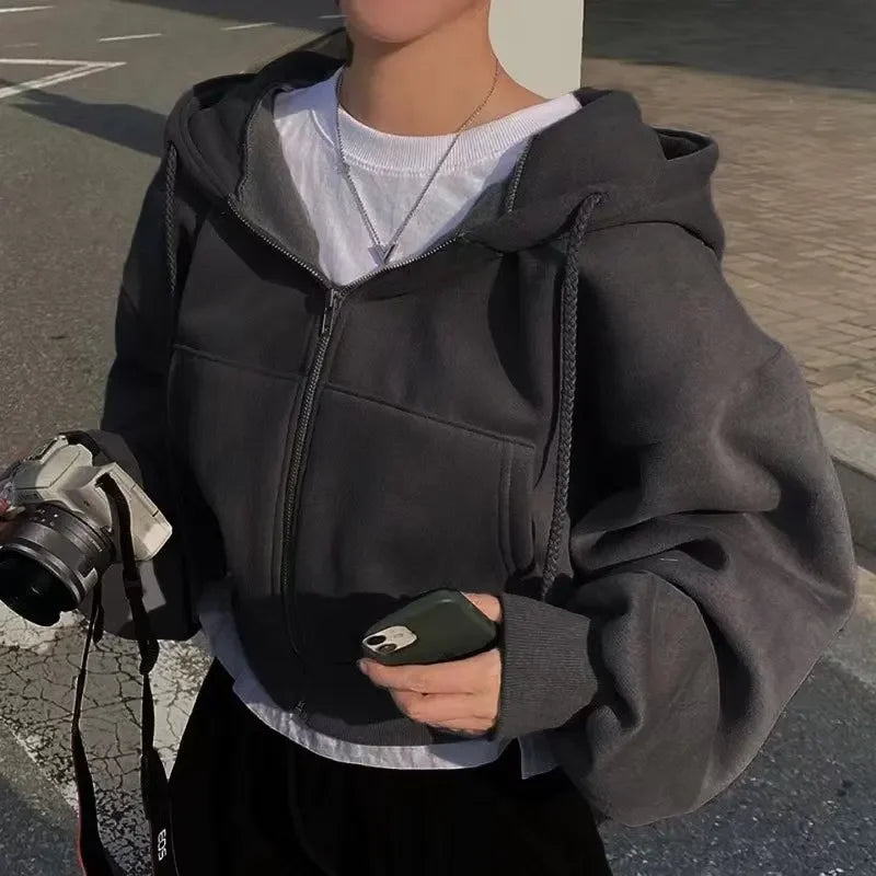 Une femme porte un sweat à capuche gris, avec une coupe ample et décontractée. Elle tient un appareil photo dans une main et un téléphone portable dans l'autre. Le sweat est zippé et elle porte un t-shirt blanc dessous. Elle se trouve en extérieur, avec un arrière-plan urbain. La lumière du soleil éclaire doucement l'ensemble de la scène.