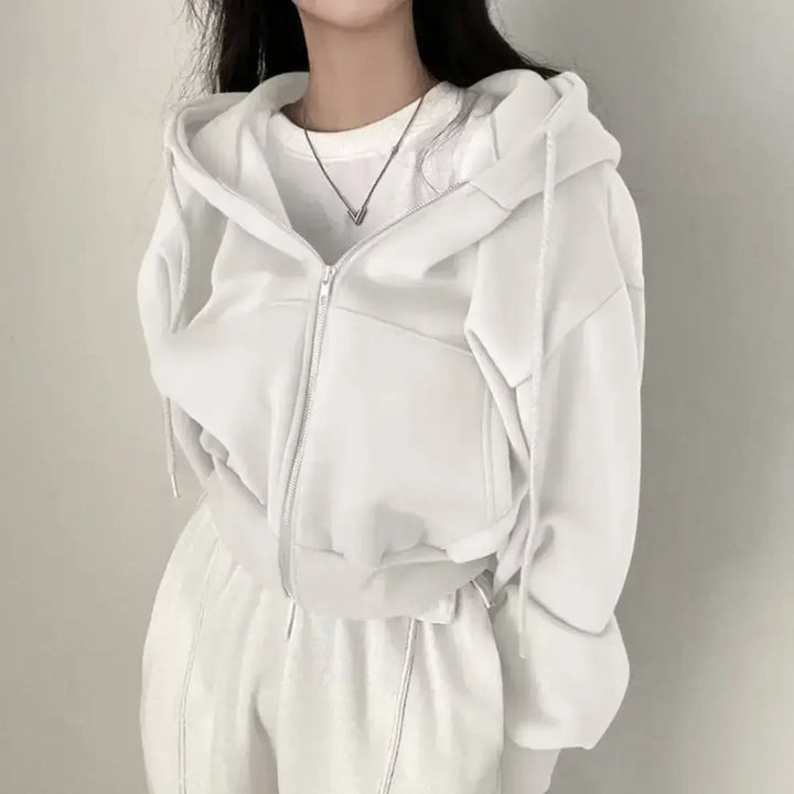 Une femme porte un sweat à capuche blanc, avec une coupe ample et décontractée. Le sweat est zippé et elle porte également un t-shirt blanc dessous. Elle a une chaîne fine autour du cou. Elle se tient de face, montrant l'ensemble du look avec une ambiance minimaliste et moderne.