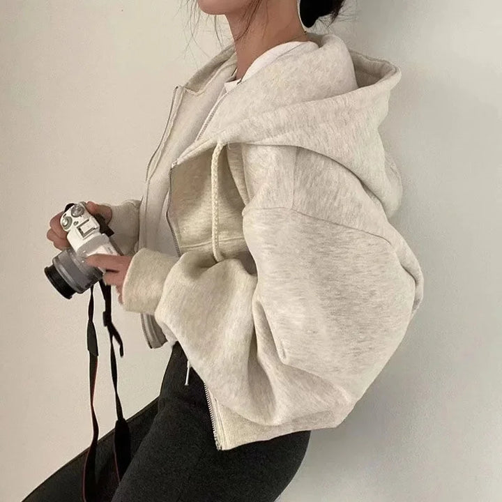 Une femme porte un sweat à capuche beige avec des manches volumineuses. Elle tient un appareil photo en main, et son look décontracté est complété par un pantalon noir. Le sweat est zippé et ajusté au niveau du bas.