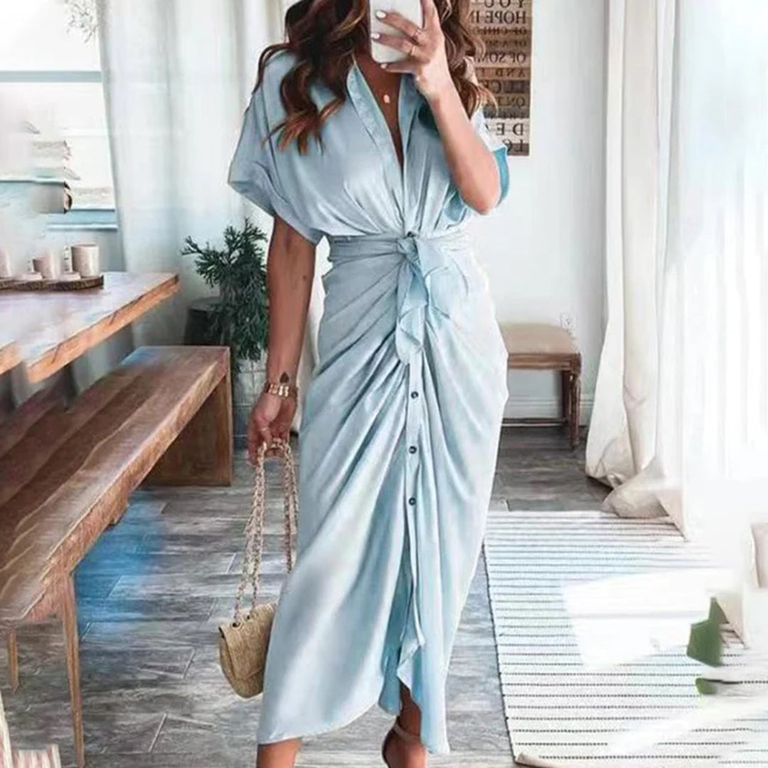 Robe midi bleu ciel avec une coupe fluide et une taille nouée, créant une silhouette élégante. Les boutons sur le devant ajoutent une touche moderne et raffinée, tandis que les accessoires dorés et le sac à main complètent le look chic, parfait pour des occasions décontractées ou formelles.