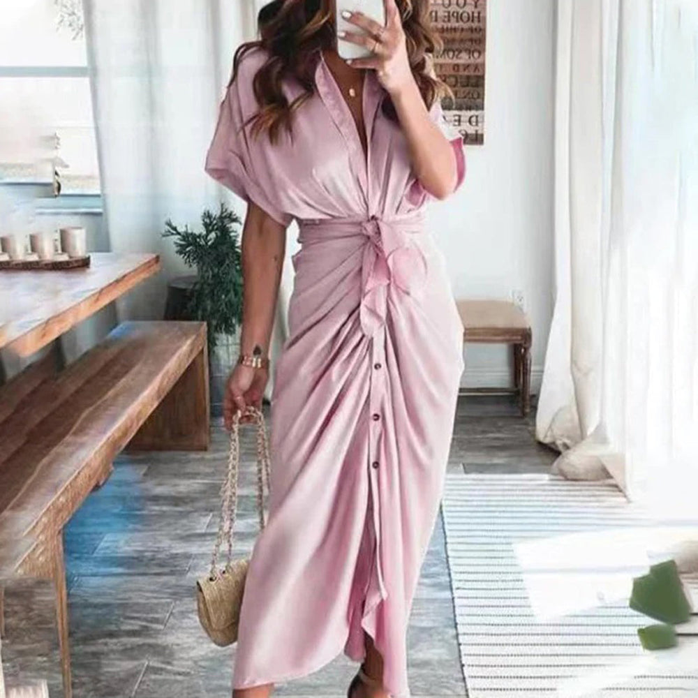 Robe midi rose délicate avec une coupe fluide et une taille nouée, soulignant la silhouette féminine. Les boutons sur le devant ajoutent un détail élégant et moderne, tandis que les accessoires dorés et les talons complètent le look chic, parfait pour des événements ou des occasions spéciales.