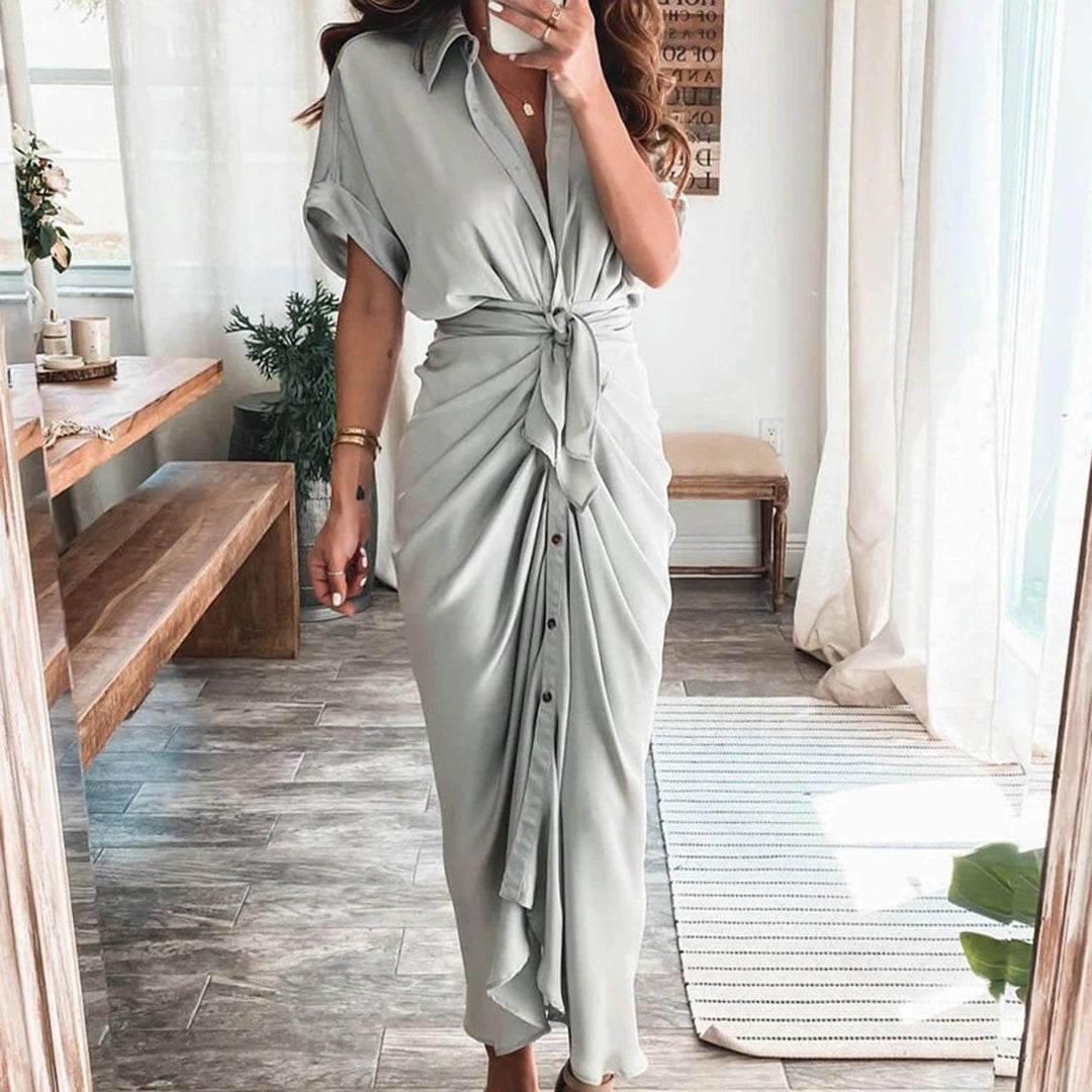 Robe midi gris clair avec une coupe fluide et une taille nouée, créant une silhouette élégante. Les boutons sur le devant apportent une touche moderne et chic, tandis que les accessoires dorés et les sandales à talons ajoutent une touche sophistiquée à ce look parfait pour les occasions spéciales.