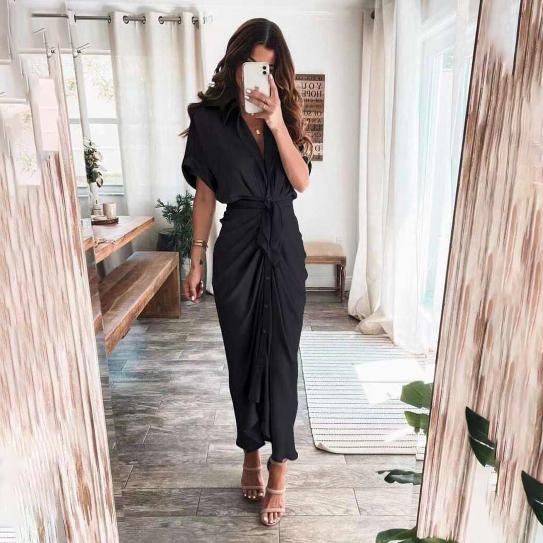 Robe midi noire avec une coupe fluide et une taille nouée, accentuant la silhouette. Les boutons sur le devant ajoutent une touche élégante, tandis que les sandales à talons et l'ambiance lumineuse créent un look chic et moderne, idéal pour les occasions spéciales ou les sorties élégantes.
