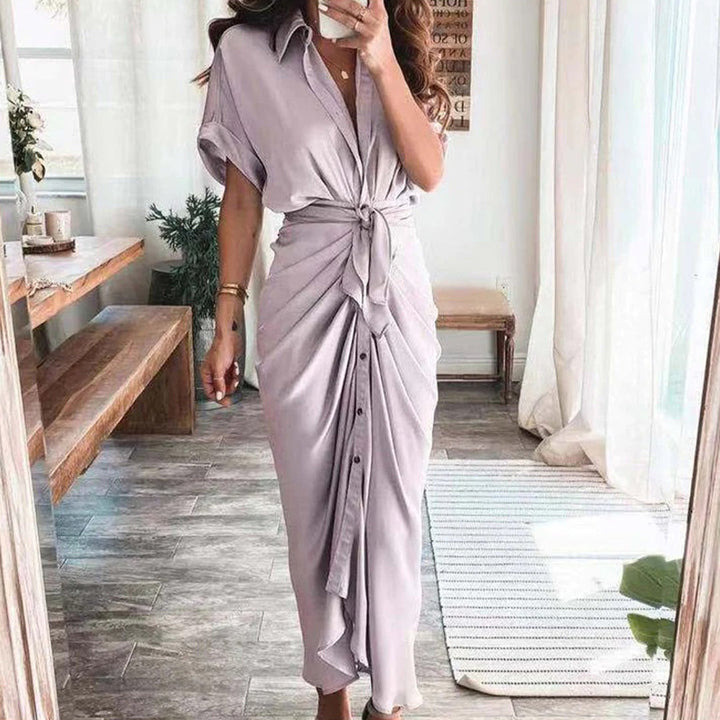 Robe midi lavande avec une coupe fluide et une taille nouée, créant une silhouette élégante et féminine. Les boutons sur le devant ajoutent un détail raffiné, tandis que le look est complété par des accessoires dorés et des sandales à talons, parfait pour des occasions décontractées ou formelles.