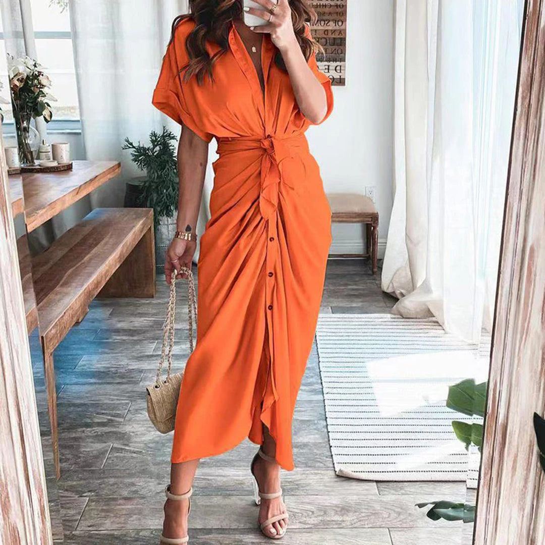 Robe midi orange vif avec une coupe fluide et une taille nouée, soulignant la silhouette. Les boutons sur le devant ajoutent une touche élégante, tandis que les sandales à talons et le sac à main créent un look moderne et chic, parfait pour les occasions décontractées ou estivales.