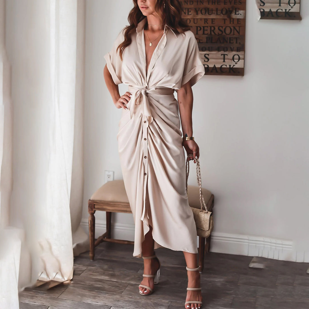 Robe midi beige élégante avec une coupe cintrée et une taille nouée, mettant en valeur une silhouette féminine. Les boutons sur le devant ajoutent une touche de style moderne, tandis que la pochette et les sandales à talons complètent le look chic et raffiné, parfait pour des événements ou des sorties décontractées.