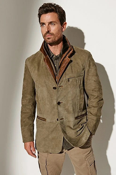 Un homme porte une veste en daim marron clair avec des finitions en cuir marron foncé au col et aux poches. Le blazer présente une coupe structurée à deux boutons et un col à revers contrastant, associé à un pantalon cargo beige. L’homme adopte une posture confiante dans un éclairage doux mettant en valeur la texture luxueuse du daim.