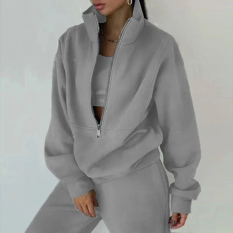 Une femme porte un ensemble décontracté gris clair composé d'un haut à zip et d'un pantalon assorti. Le haut est légèrement oversize avec un col zippé et elle a l'air détendue et élégante. Le fond est neutre et minimaliste.