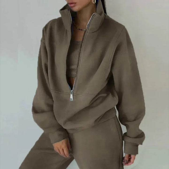 Une femme porte un ensemble décontracté de couleur marron, comprenant un haut à zip et un pantalon assorti. Le haut à col zippé lui donne un look moderne et confortable, parfait pour un style décontracté pendant la saison hivernale.