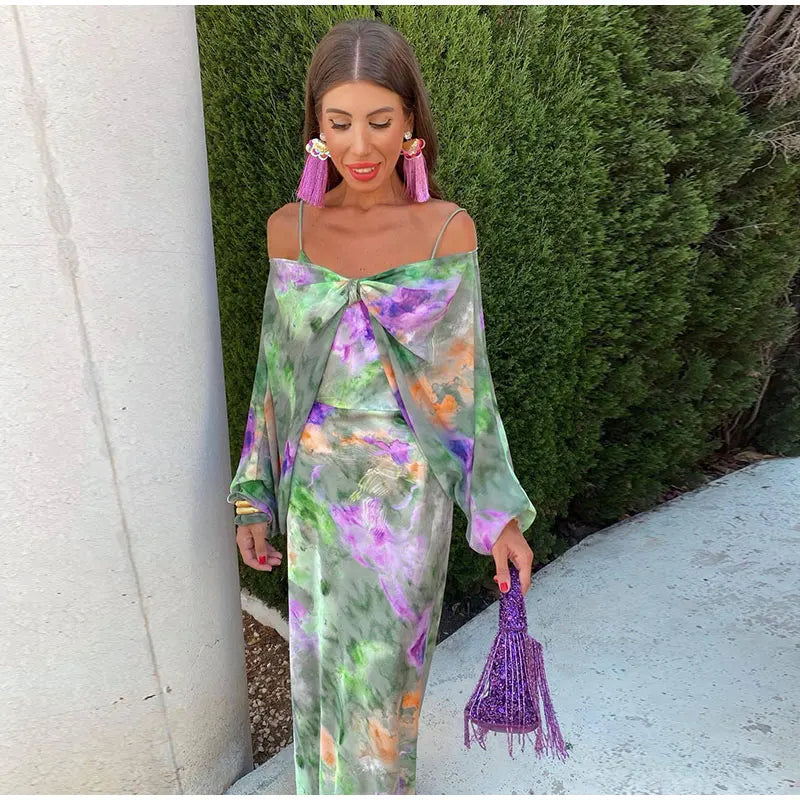 Robe maxi de soirée Kyra pour femme, élégante et fluide, à imprimé floral vert et violet avec épaules dénudées et détail noué sur le devant. Style chic et tendance, accompagnée d’accessoires dorés et d’une pochette violette, parfaite pour une tenue de soirée raffinée.