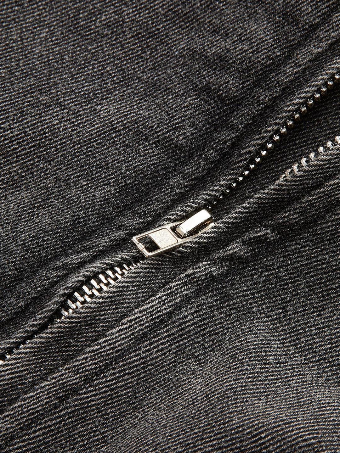 Gros plan sur le détail de la fermeture zippée en métal argenté sur denim gris délavé, mettant en valeur la finition soignée et le style moderne de la veste.