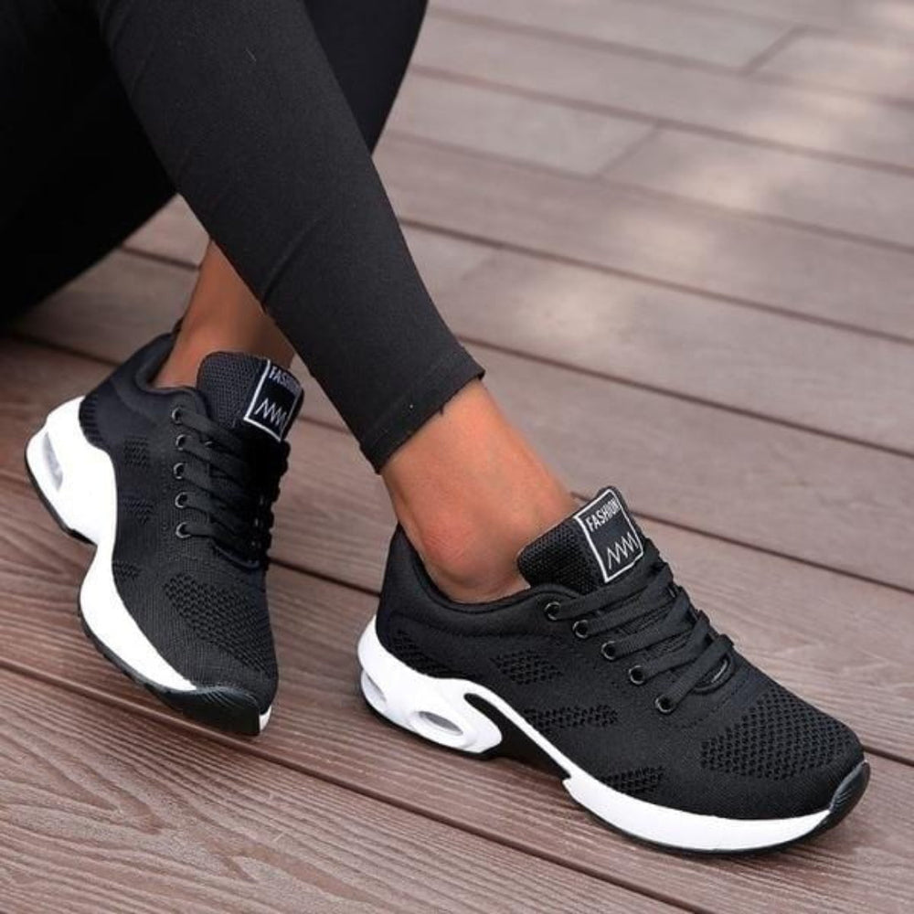 Chaussures de sport basses pour femme, de couleur noire avec semelle blanche épaisse, portées avec un legging noir, au design moderne et respirant, idéales pour un usage quotidien.
