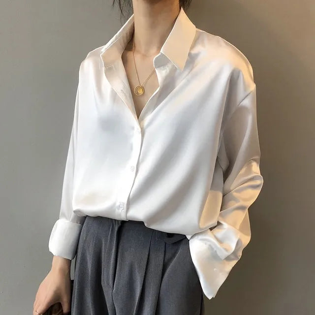 Femme portant une blouse blanche fluide au style tendance, avec col chemise, manches longues légèrement retroussées et coupe ample, associée à un pantalon taille haute, offrant une allure élégante et moderne.