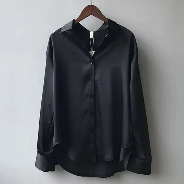 Blouse noire fluide suspendue sur cintre, au style tendance, avec col chemise, fermeture boutonnée sur le devant et manches longues à poignets larges, offrant une coupe élégante et moderne.