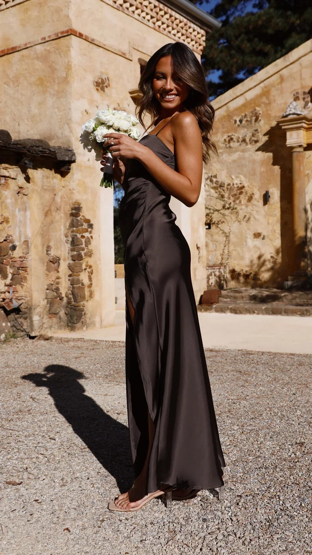 Robe maxi Leona pour femme, dans une teinte noire élégante, avec des bretelles spaghetti fines et une fente latérale sensuelle. La robe fluide et sophistiquée met en valeur la silhouette et est idéale pour des événements spéciaux. Le modèle porte la robe avec un bouquet de fleurs blanches, créant une ambiance chic et lumineuse, parfaite pour des occasions formelles ou estivales.