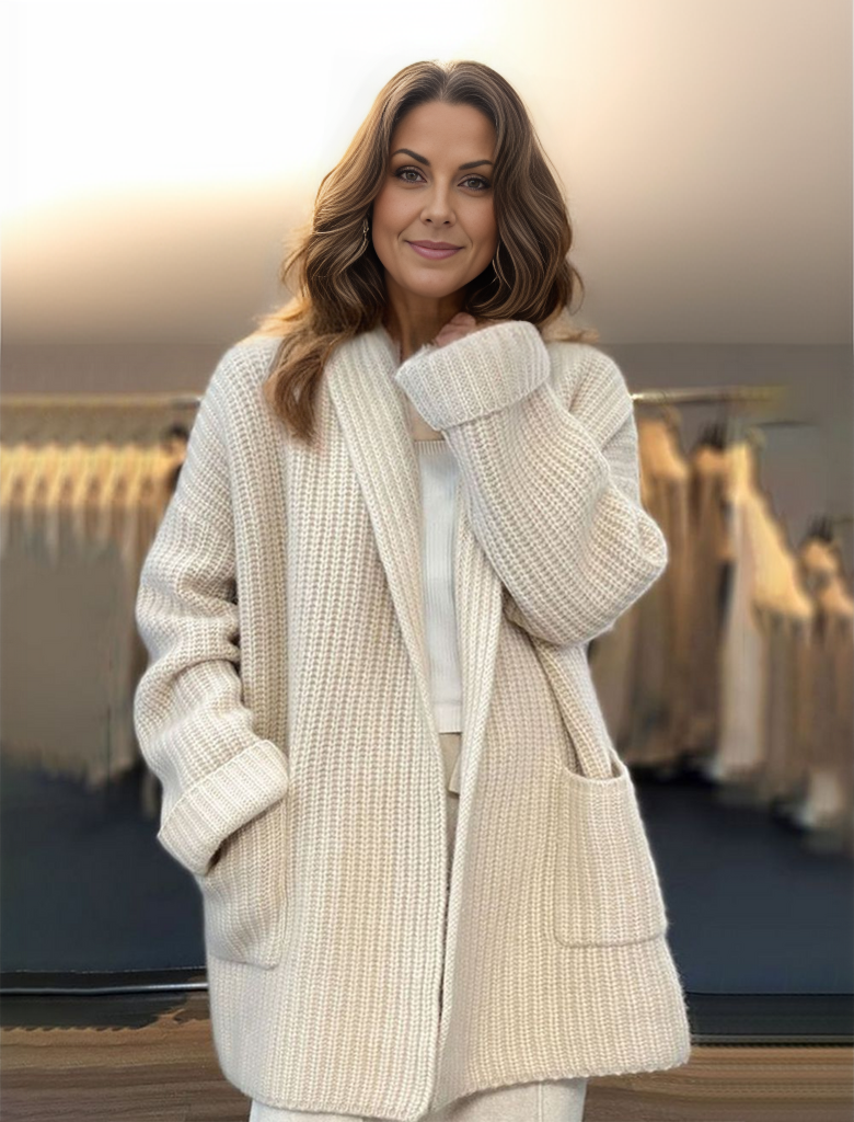 Une femme souriante, vêtue d'un long cardigan épais de couleur beige, dans un style moderne et confortable, se tient dans un intérieur chaleureux avec des vêtements duveteux en arrière-plan.