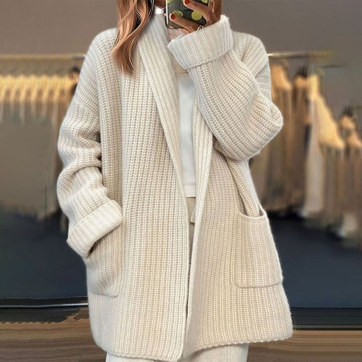 Femme prenant une photo dans un miroir, vêtue d'un long cardigan épais en maille beige, avec des poches avant et un style décontracté, dans un intérieur aux tons doux.