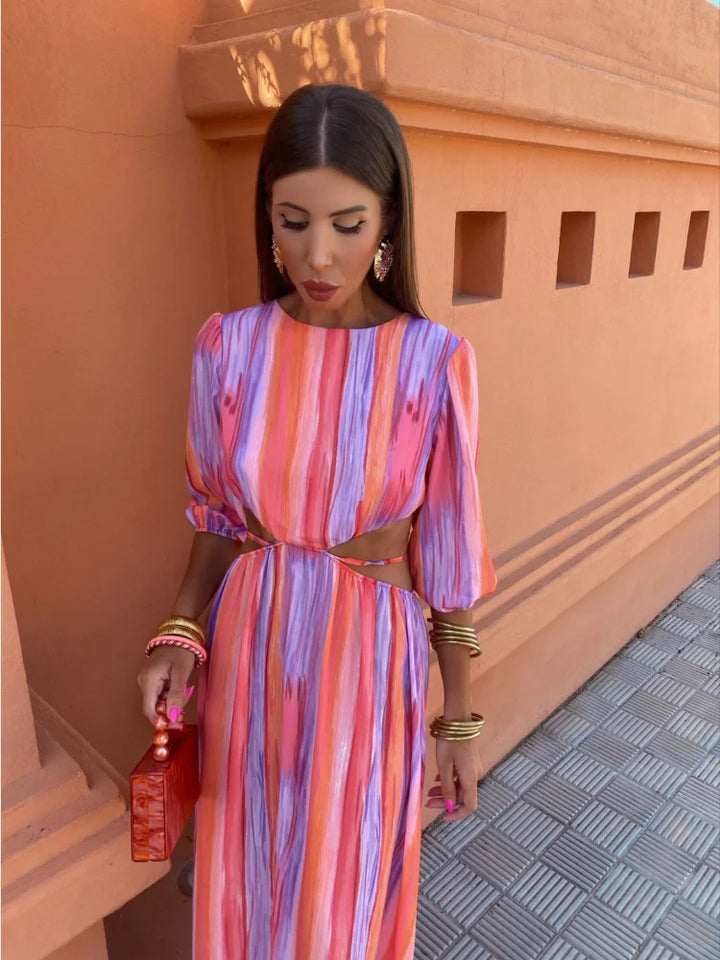 Robe longue aux motifs tie-dye vibrants dans des tons de rose, violet et orange, avec des découpes élégantes sur les côtés. Accessoirisée avec un sac à main rouge et des bijoux dorés, cette robe incarne un style audacieux et moderne, idéale pour les occasions estivales.