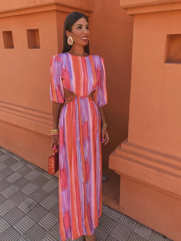 Robe longue aux motifs tie-dye vibrants dans des tons de rose, violet et orange, avec des découpes stylisées sur les côtés. Cette robe, parfaite pour les occasions estivales et les sorties tendance, est complétée par un sac à main rouge et des accessoires dorés, créant une silhouette chic et moderne.