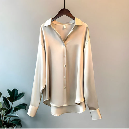 Une blouse satinée chic pour femme en beige clair, avec un col classique, des manches longues avec poignets à boutons et une fermeture à boutons sur le devant. La blouse présente une finition lisse et élégante, suspendue à un cintre en bois contre un fond neutre avec une plante au premier plan.
