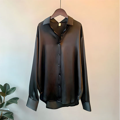 Une blouse satinée chic pour femme en noir, avec un col classique, des manches longues avec poignets à boutons et une fermeture à boutons sur le devant. La blouse a une finition lisse et brillante, suspendue à un cintre en bois contre un fond doux et neutre, avec une plante au premier plan.