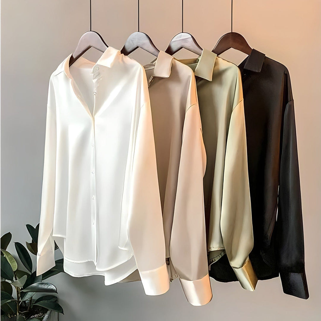 Une collection de quatre blouses satinées pour femme dans différentes couleurs (blanc, beige, olive et noir), accrochées à des cintres en bois. Chaque blouse présente un col classique, des manches longues avec poignets à boutons et une finition satinée lisse et brillante, exposée sur un fond neutre avec une plante au premier plan.