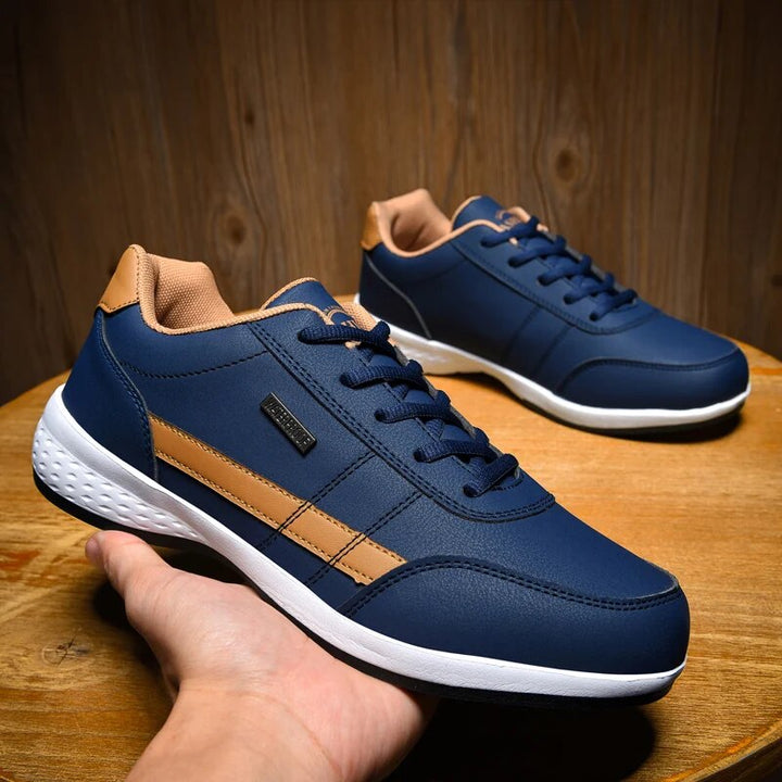 Chaussures décontractées pour homme Loïc modernes, baskets bleu marine avec détails camel et semelle blanche, présentées sur table en bois avec une paire en arrière-plan.