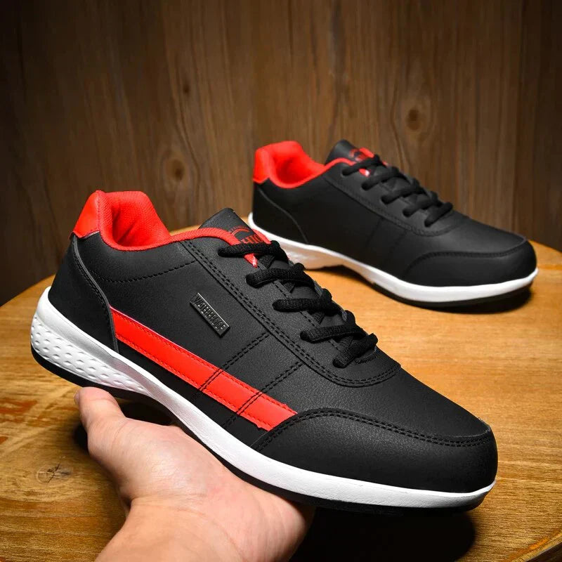 Chaussures décontractées pour homme Loïc modernes, baskets noires avec bande rouge et col rouge, lacets noirs et semelle blanche, présentées sur table en bois avec une paire en arrière-plan.