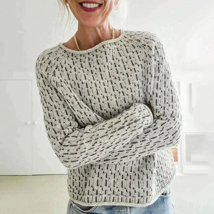 Femme souriante portant un pull à col rond en maille texturée gris clair, avec manches longues et coupe décontractée, posant dans une pièce lumineuse au style minimaliste.