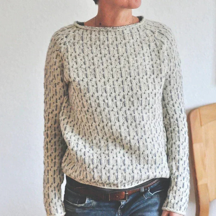 Femme portant un pull à col rond en maille texturée gris clair, avec manches longues et coupe décontractée, associant le vêtement à un jean foncé dans un intérieur simple et lumineux.