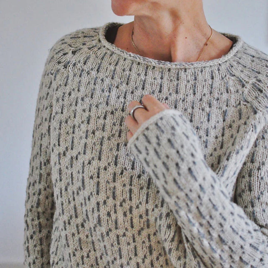 Gros plan sur une femme portant un pull à col rond en maille texturée gris clair, mettant en valeur la douceur du tissu et les manches longues légèrement oversize, dans un intérieur lumineux.