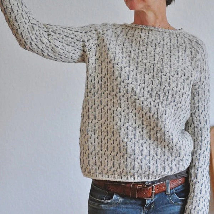 Femme portant un pull à col rond en maille texturée gris clair, avec manches longues et coupe confortable, associé à un jean et un ceinture en cuir brun, posant dans un intérieur lumineux.