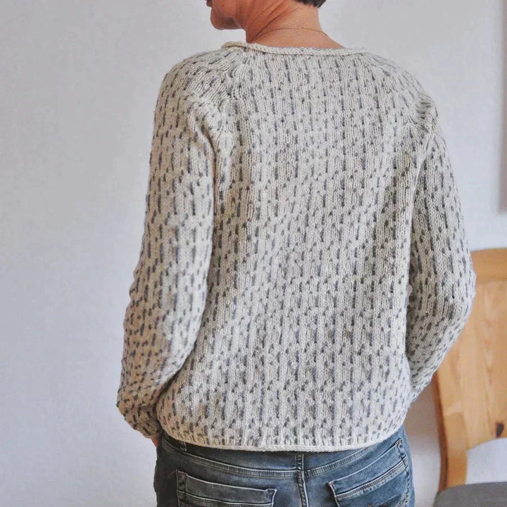 Vue de dos d’une femme portant un pull à col rond en maille texturée gris clair, avec manches longues et coupe confortable, assorti à un jean dans un intérieur lumineux.