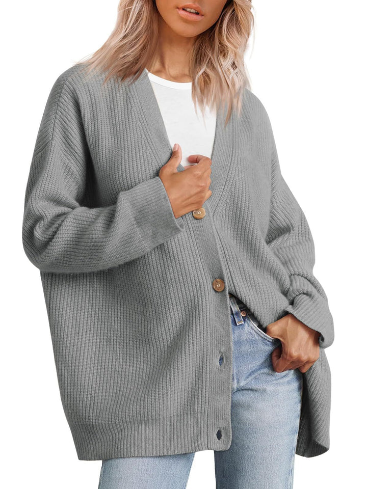 Femme portant un cardigan gris à col en V, doté de gros boutons en bois et d’une coupe oversize. Le vêtement est superposé à un t-shirt blanc et associé à un jean clair, offrant un style moderne, décontracté et confortable.