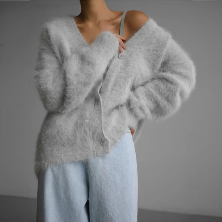 Femme portant un cardigan ample et duveteux de couleur gris clair, légèrement tombant sur l’épaule, associé à un pantalon large de teinte pastel. Le style évoque une allure douce, confortable et élégante, parfaite pour un look cocooning moderne.