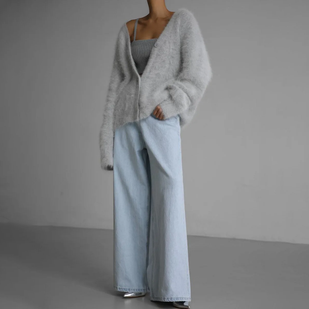 Femme vêtue d’un cardigan duveteux gris clair légèrement oversize, porté sur un débardeur côtelé gris, associé à un jean large bleu pâle et des chaussures argentées. Le look dégage une allure décontractée et moderne, alliant douceur, confort et élégance minimaliste.