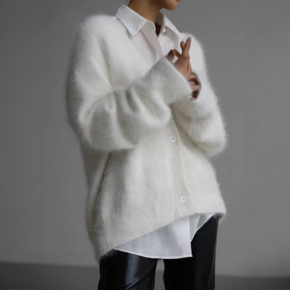 Une femme porte un cardigan blanc ample et duveteux sur un tailleur blanc élégant, associé à un pantalon noir en similicuir. L'ensemble crée un contraste raffiné entre douceur et modernité, idéal pour un look chic et minimaliste.