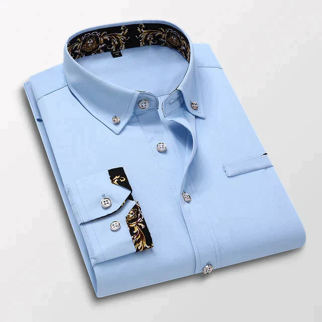 Chemise longue pour hommes Ludovic moderne bleu clair, pliée avec col boutonné et détails baroques dorés à l’intérieur du col et sur les poignets, présentée sur fond clair.